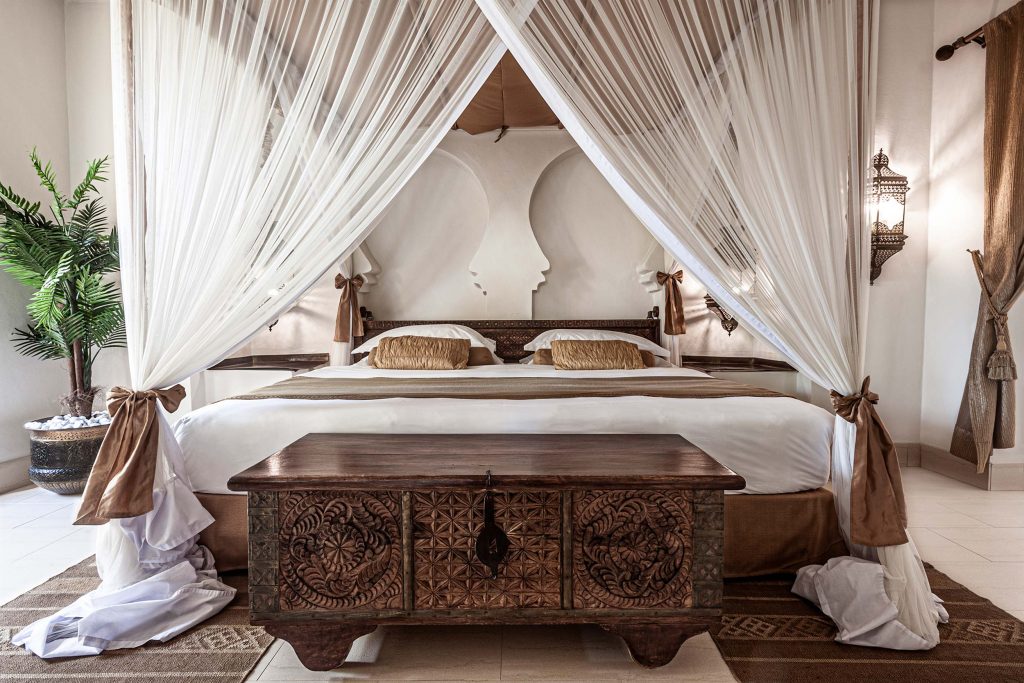 Hotelzimmer mit großem Himmelbett, weißen Vorhängen, Holztruhe und metallischen Wandleuchten im Baraza Resort & Spa