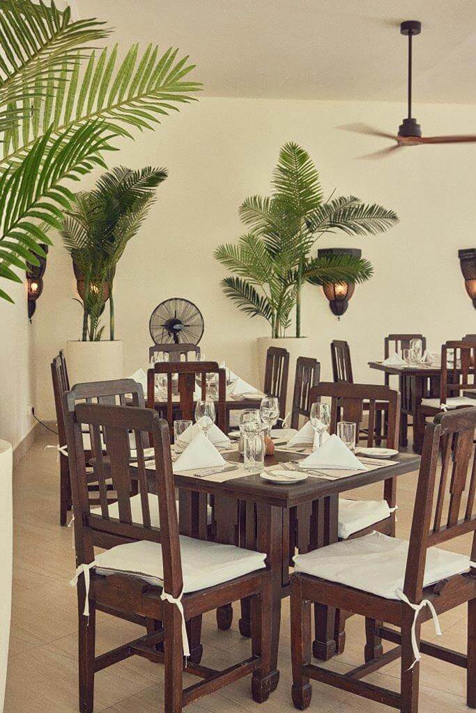 Helles Hotelrestaurant mit dunklen Holztischen, weißen Servietten, Gläsern, weißen Stühlen und grünen Pflanzen in Sansibar