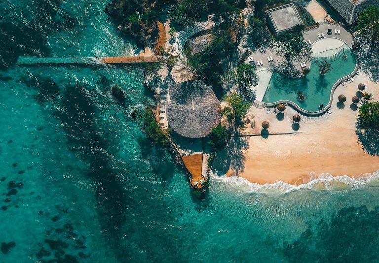 Vogelperspektive auf Bawe Island Resort mit Pool, weißen Liegestühlen, Strand und hölzernem Steg