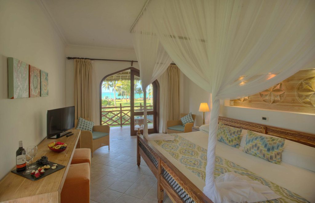 Hotelzimmer mit Himmelbett aus Holz, Tisch mit Wein und Obst, Blick auf Palmen und Meer in Sansibar