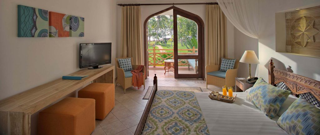Modernes Hotelzimmer mit Doppelbett, Holztisch, orangenen Hockern und bogenförmiger Glastür mit Blick auf tropische Pflanzen