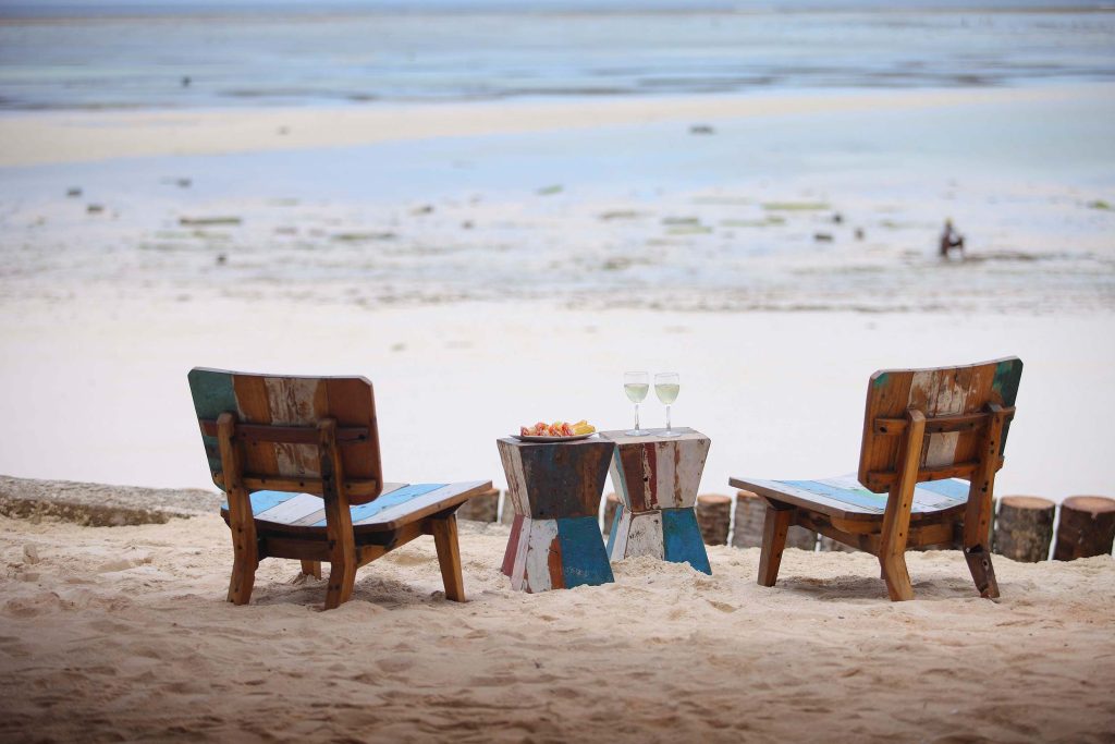 Zwei bunte Holzsesseln mit Holztisch auf Sandstrand und Meer im Hintergrund bei Tageslicht in Sansibar