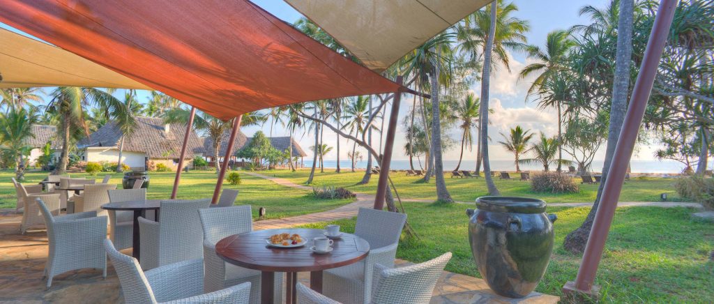 Außenbereich mit weißen geflochtenen Stühlen, Holztisch und bunten Segeltuchüberdachungen im Bluebay Beach Resort Sansibar