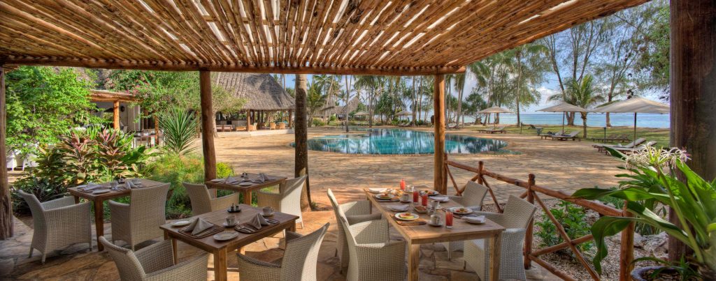 Überdachte Terrasse mit Holzbalken, Rattanstühlen und Tischen, Blick auf Pool und Liegestühle in Sansibar