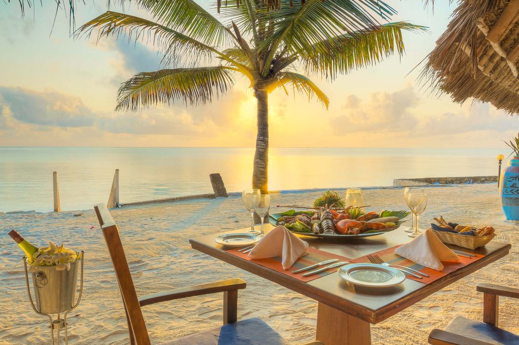 Holztisch mit Meeresfrüchten, Wein und Gläsern am Strand mit Palme und Meer bei Sonnenuntergang
