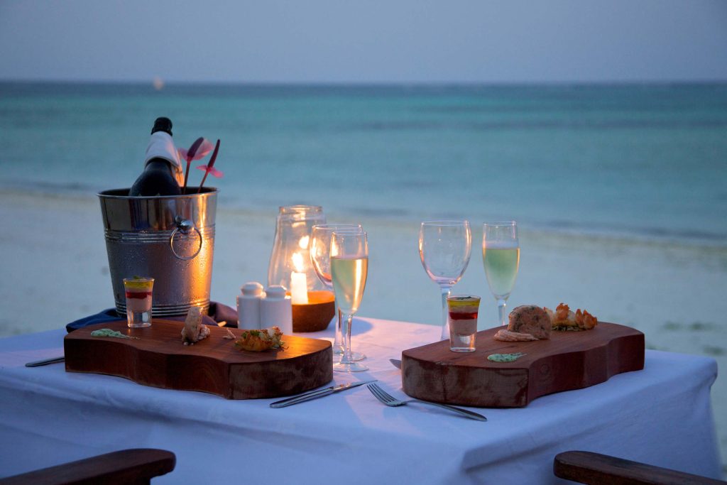Abendessen am Strand mit Tisch, weißen Tüchern, Holzplatten mit Häppchen, Weingläsern und Meer im Hintergrund