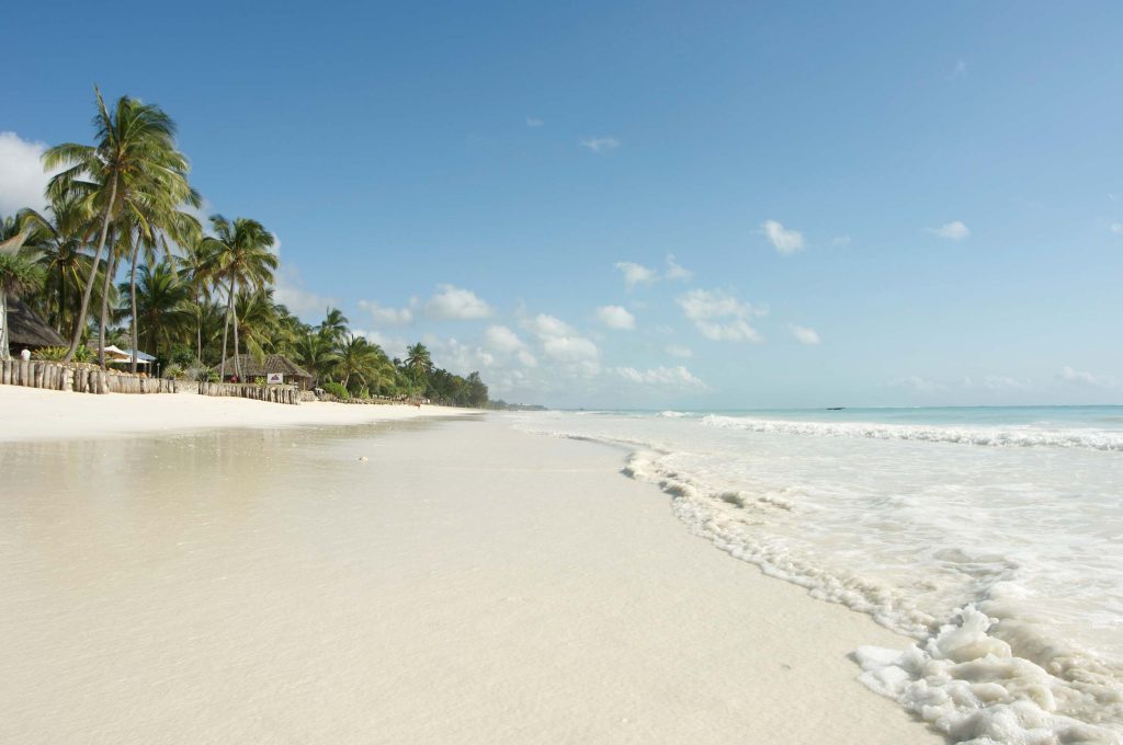 weiter weißer Sandstrand mit Palmen, Holzhütten im Hintergrund, türkisblaues Meer und blauer Himmel