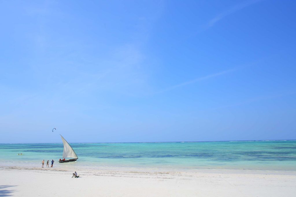 Breiter Sandstrand mit türkisfarbenem Meer und traditionellem Segelboot bei klarem blauem Himmel