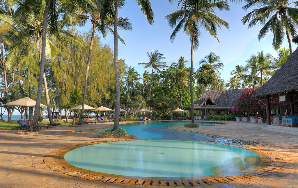 Großer Swimmingpool mit hohen Palmen und strohgedeckten Gebäuden im Bluebay Beach Resort & Spa auf Sansibar
