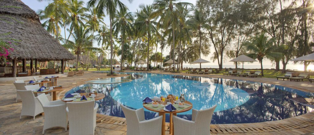 Hotelpool mit Palmen, Liegestühlen und Sonnenschirmen, davor Holztische mit weißen geflochtenen Stühlen und gedecktem.