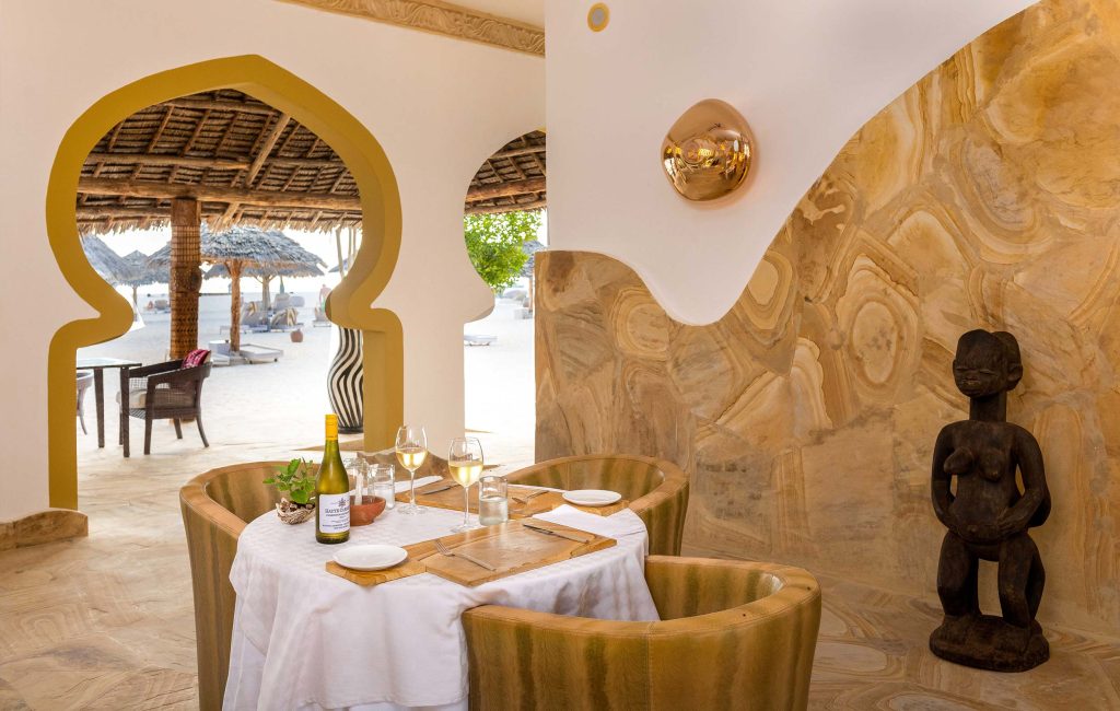 Hotelzimmer mit gedecktem Tisch, Rattansesseln, geschnitzter Statue und Blick auf Strandliegen
