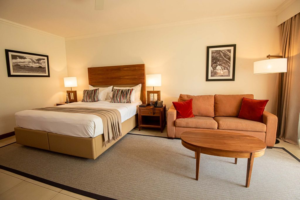 Hotelzimmer mit großem Bett, hölzernem Kopfteil, zwei Nachttischen, Sofa mit orangefarbenem Stoff und ovalem Holztisch