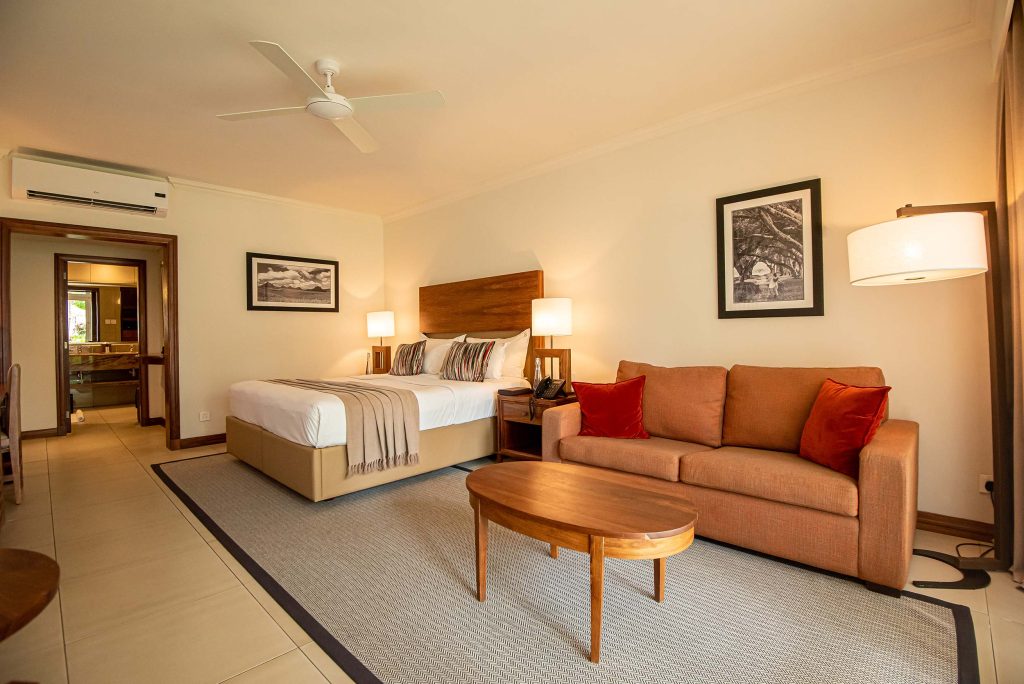 Hotelzimmer mit großem Bett, weißen Bettwäsche, beigen Überwürfen, Sofa mit roten Kissen und schwarzen Fotografien.