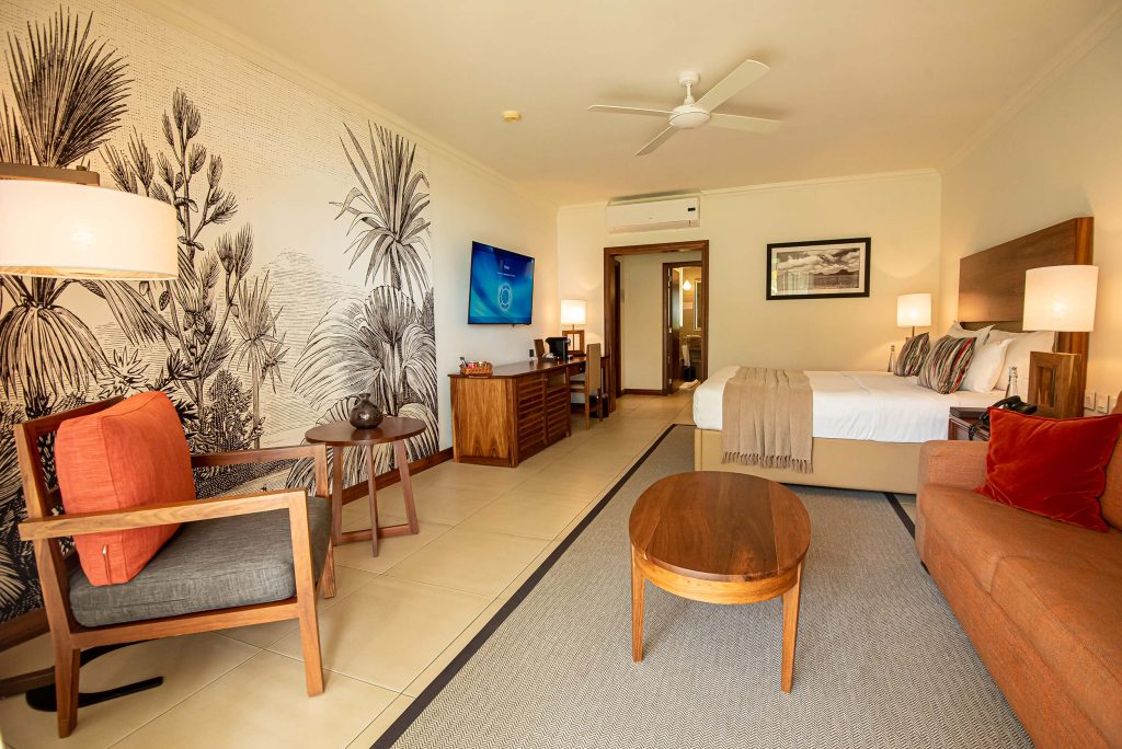 Hotelzimmer mit Doppelbett, Sofa, Sessel, Holztisch und botanischer schwarz-weißer Wandtapete in beige gehaltenen Wänden