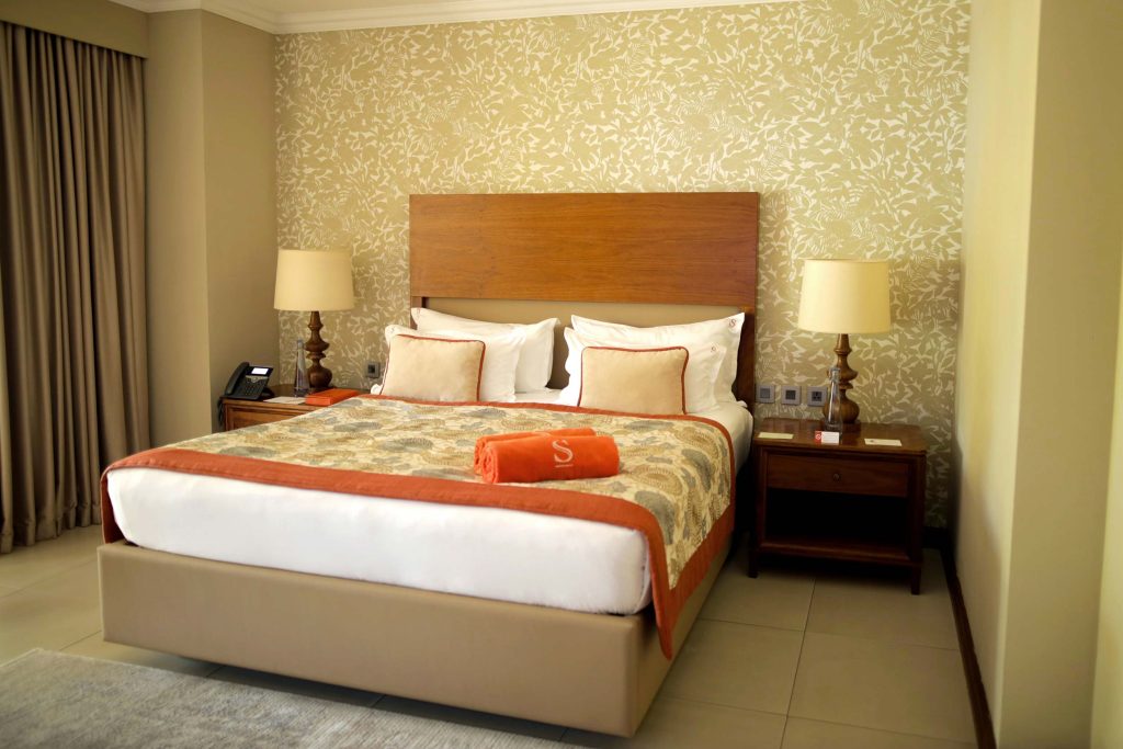 Hotelzimmer mit Doppelbett, gemusterter Bettdecke, floraler Tapete, zwei Holznachttischen und klassischer Beleuchtung