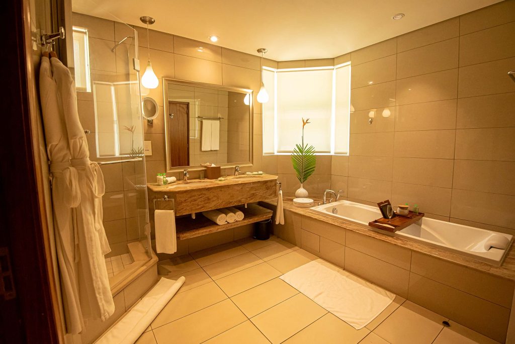 Badezimmer mit großer Badewanne, Dusche mit Glaswand, beigefarbene Fliesen und polierte Steinwaschbecken