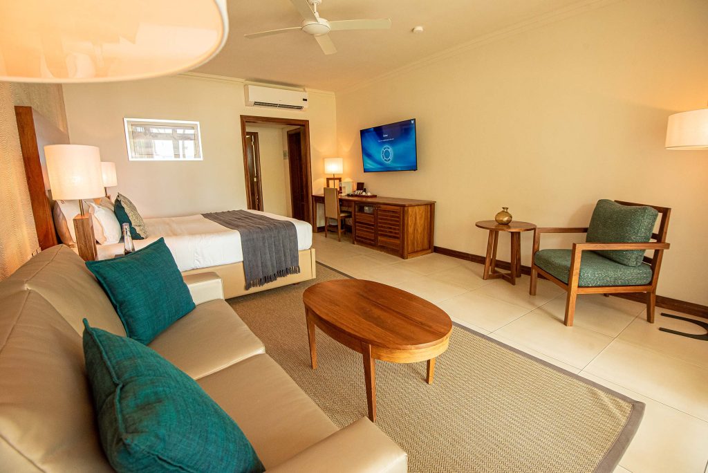 Hotelzimmer mit beigem Ledersofa, grünem Kissen, Holztisch, großem Bett mit weißen Laken und grauer Decke