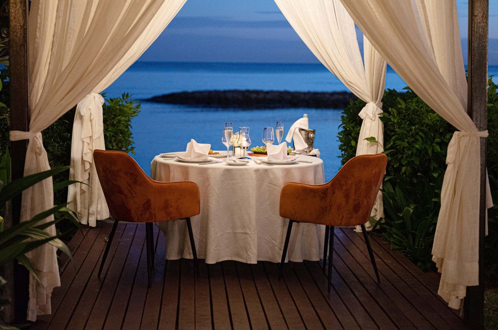 Essbereich im Freien auf Holzterrasse mit rundem Tisch, weißen Vorhängen und Blick auf das Meer in Mauritius