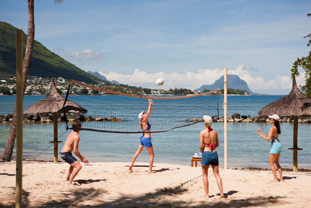 Vier Personen spielen Beachvolleyball an einem Volleyballnetz auf hellem Sandstrand mit Meer und bewaldeten Bergen.