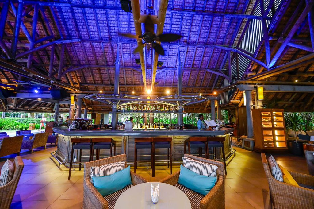 Offene Bar mit Strohdach, Bartheke aus Holz und Metall, Rattanmöbel mit bunten Kissen und grünen Pflanzen im Hintergrund