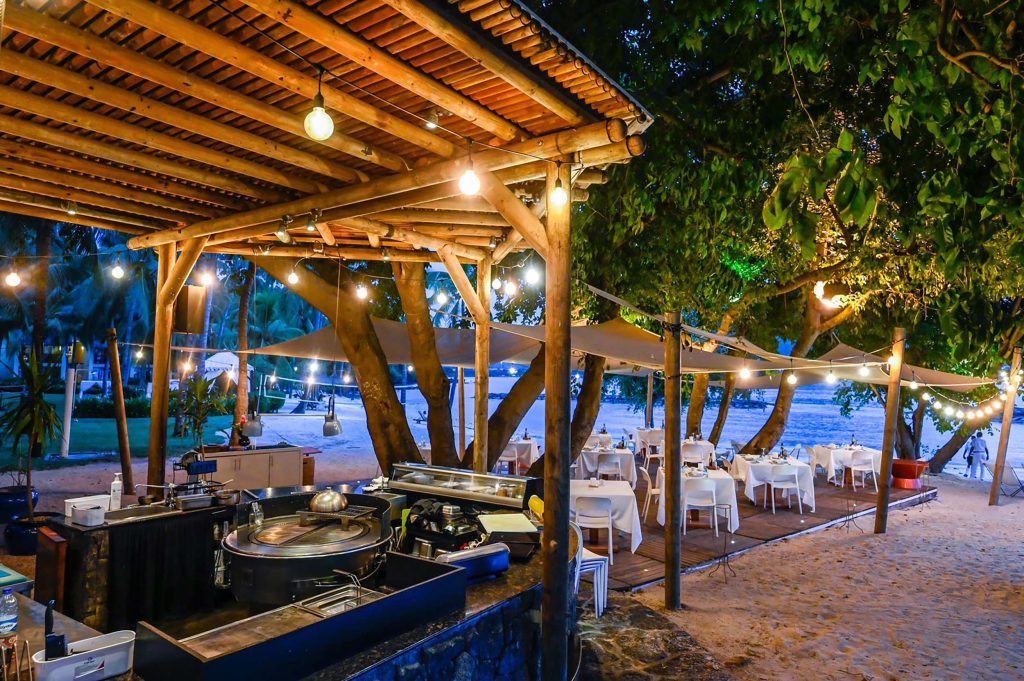 Außenbereich eines Strandrestaurants mit Holztischen, weißen Stühlen und Lichterketten auf Mauritius