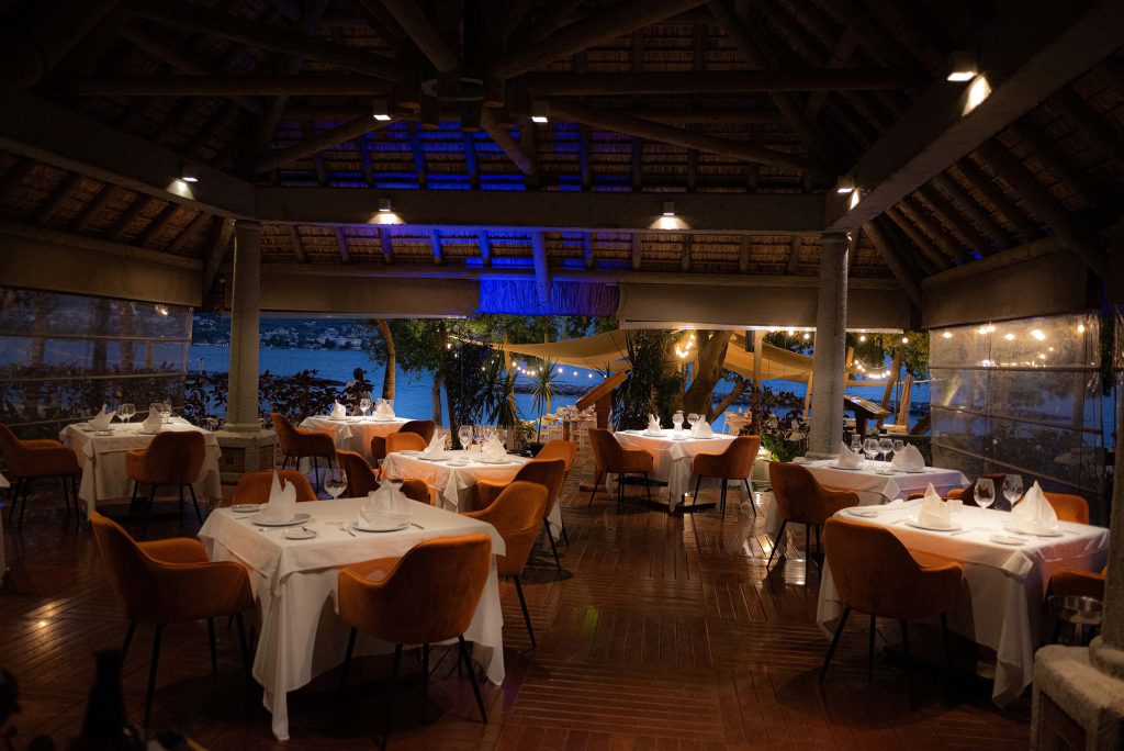Hotelrestaurant mit weißen Tischdecken, orangen Polsterstühlen, Holzdach und Blick auf Wasser