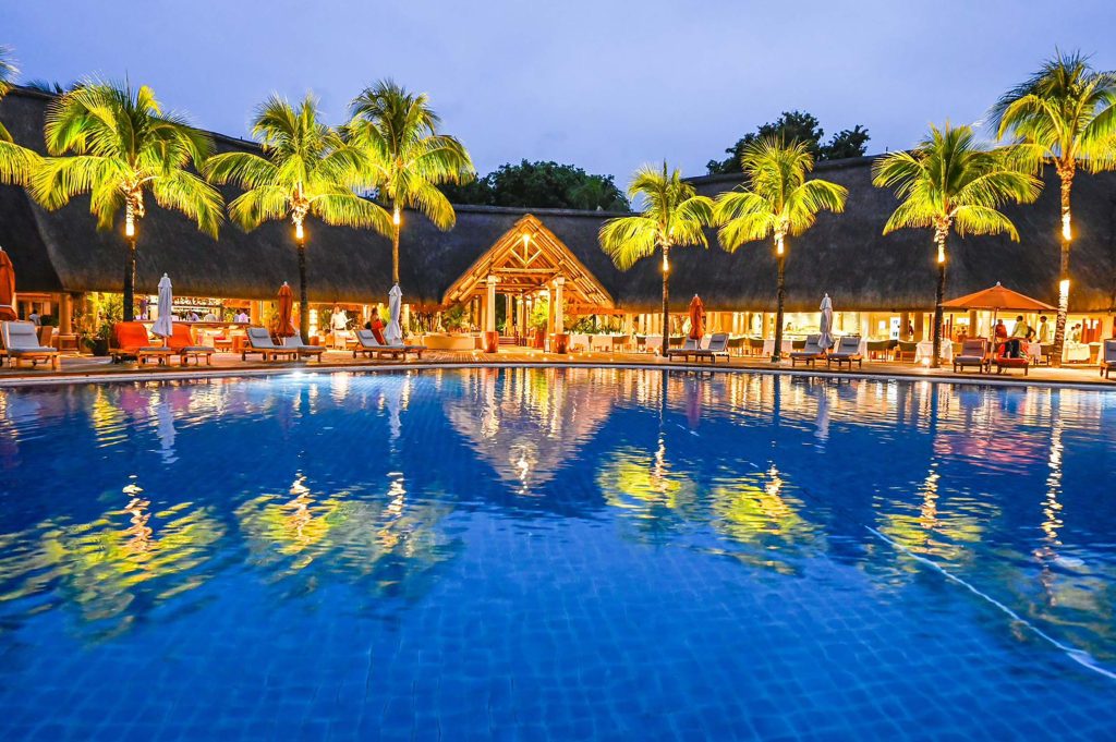 Großer Pool mit Spiegelungen von Lichtern und Palmen, Liegestühle und Gebäude mit Strohdach bei Abenddämmerung
