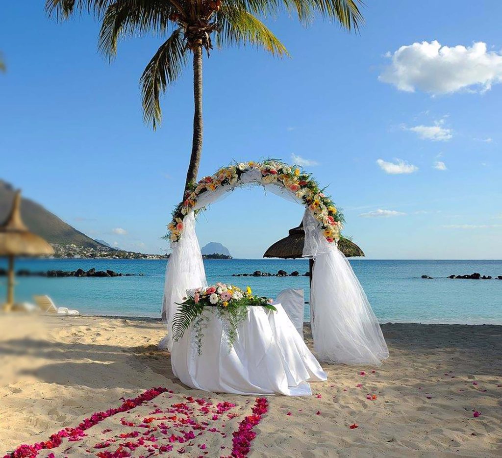 Strand mit dekoriertem Hochzeitsbogen aus weißen Tüchern und pastellfarbenen Blumen, rosa Blütenblätter auf Sand und Palme