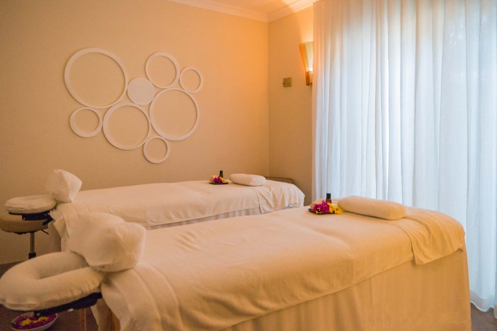 Ruhiger Spa-Raum mit zwei Massagebetten, weißen Laken, dekorativen weißen Kreisen an der Wand und großem Fenster mit hellen.