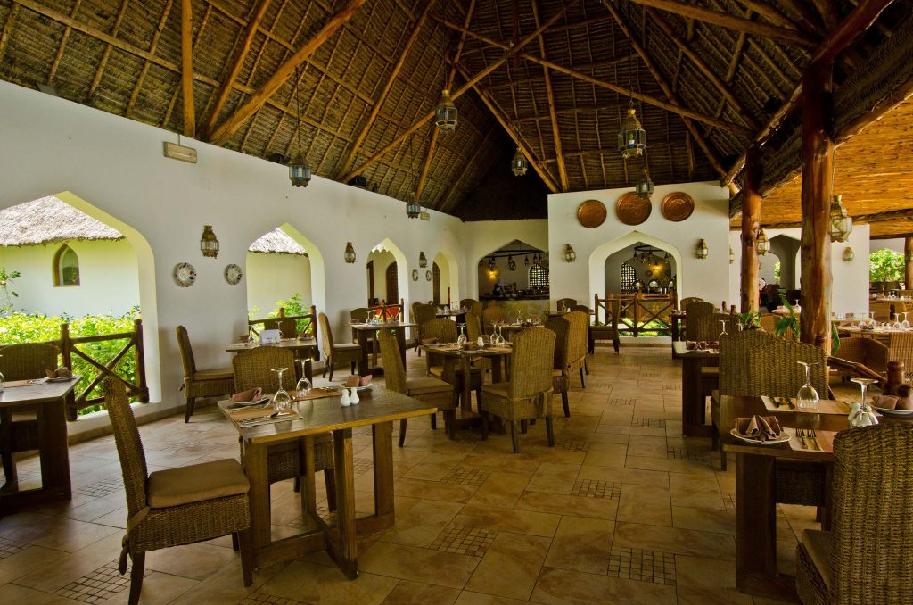 Offener Restaurantbereich mit weißen Wänden, Bögen, Holztischen, geflochtenen Stühlen und Strohdach in Sansibar