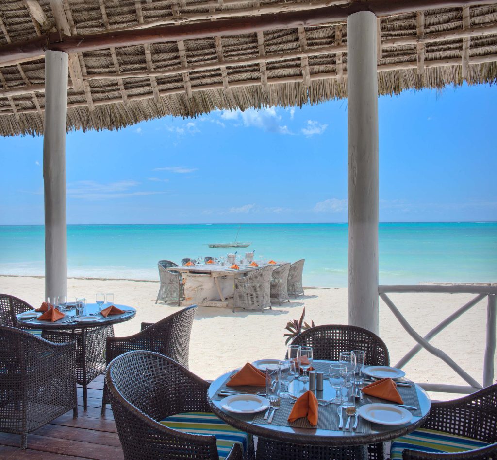 Außenbereich eines Strandrestaurants mit Palmwedeldach, runden Tischen, Rattan-Stühlen und Blick auf Meer und Sandstrand.