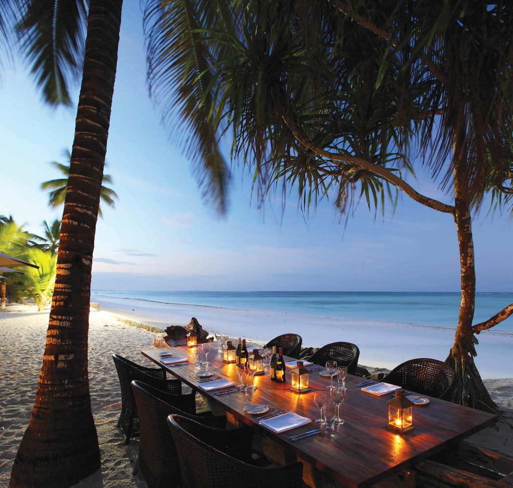 Strandrestaurant mit Holztisch, Kerzenlaternen und Rattanstühlen auf Sand, Meer und Palmen im Hintergrund