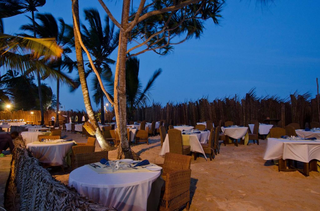 Außenbereich eines Restaurants mit runden Tischen und weißen Tischdecken, Rattanstühlen auf Sandboden, Palmen im Hintergrund