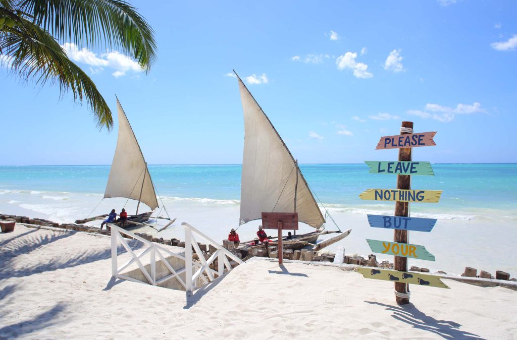 Strand mit weißem Sand, buntem Holzschild, zwei traditionellen Segelbooten und Palme am Wasser in Sansibar
