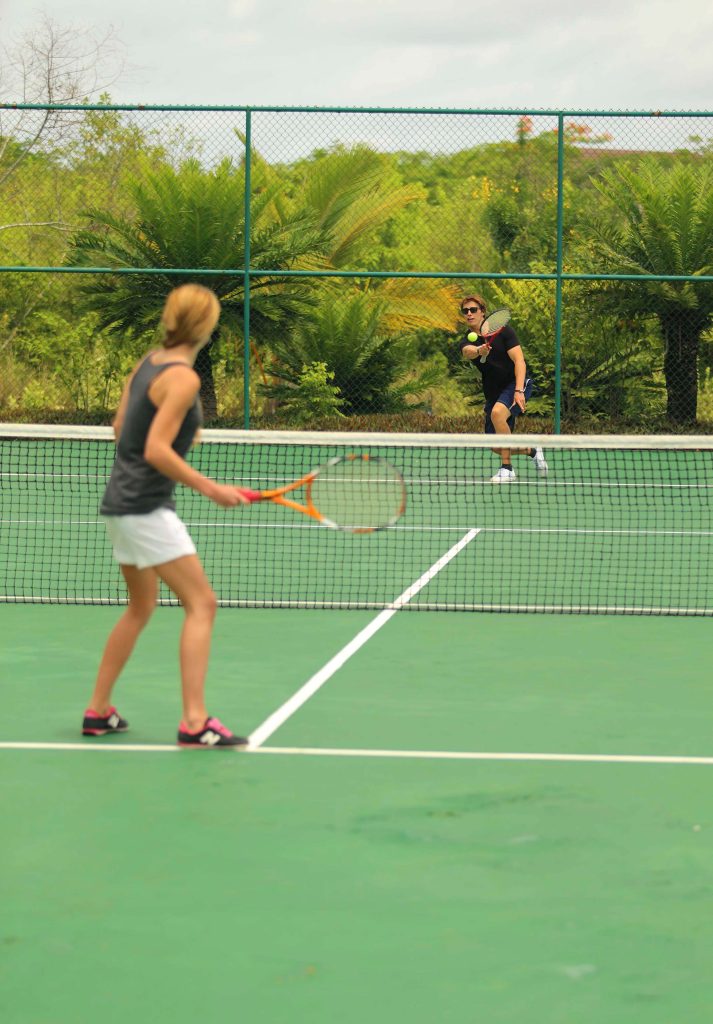 Zwei Personen spielen Tennis auf grünem Kunststoffplatz im Freien, umgeben von Palmen und Metallzaun