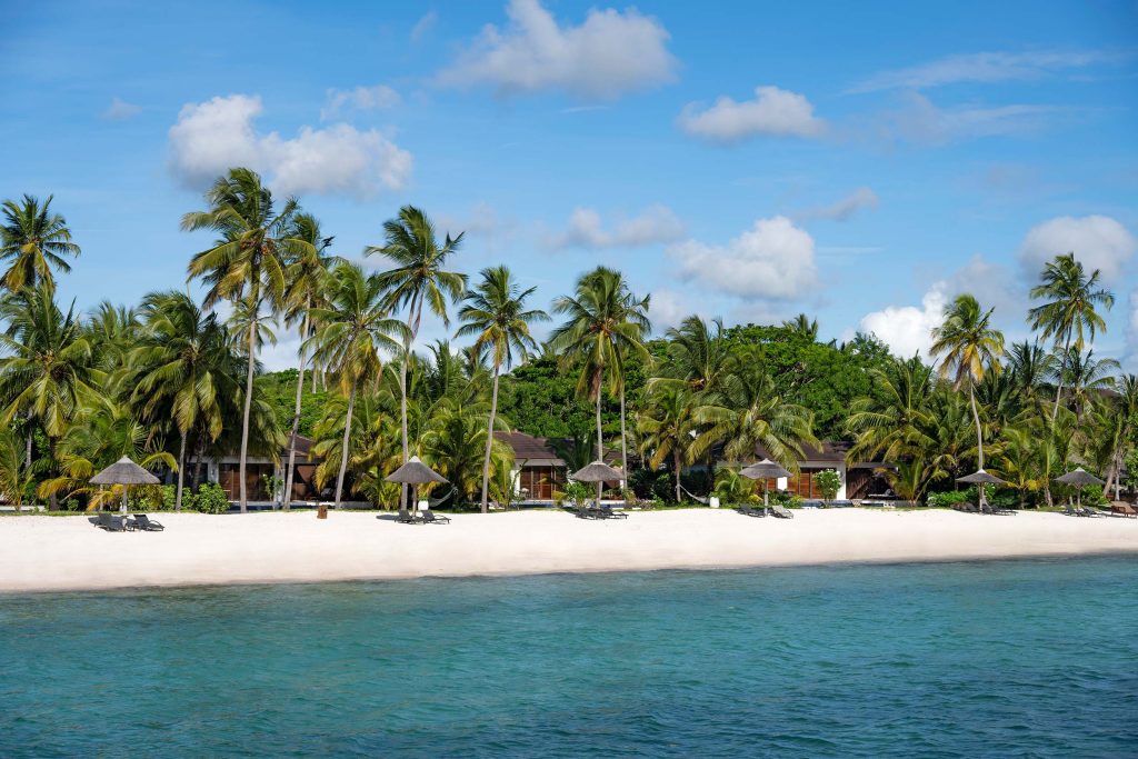 Strand mit weißem Sand, Holzziegen und Sonnenschirmen vor Palmen und ruhigem Wasser bei Tageslicht