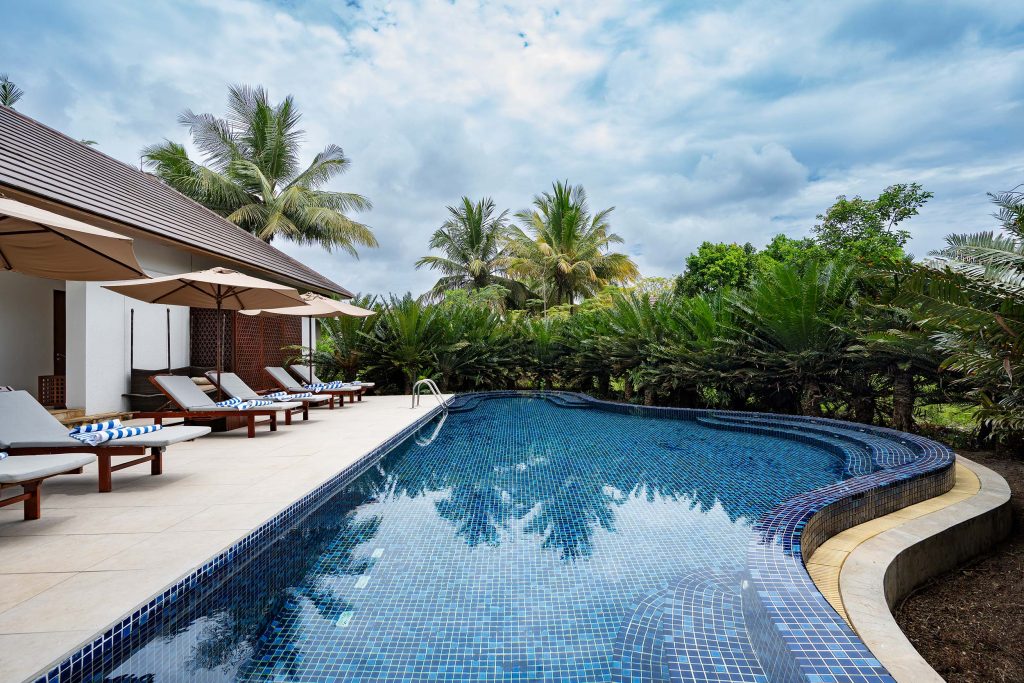 Außenbereich vom Hotel The Residence Zanzibar mit geschwungenem Pool, Holzliegen, Sonnenschirmen und tropischer Vegetation