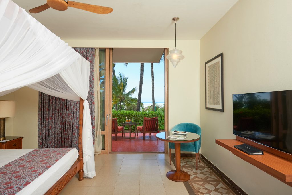 Hotelzimmer mit Himmelbett, Nachttisch, Glastüren zur Terrasse mit Stühlen und Gartenblick in Sansibar
