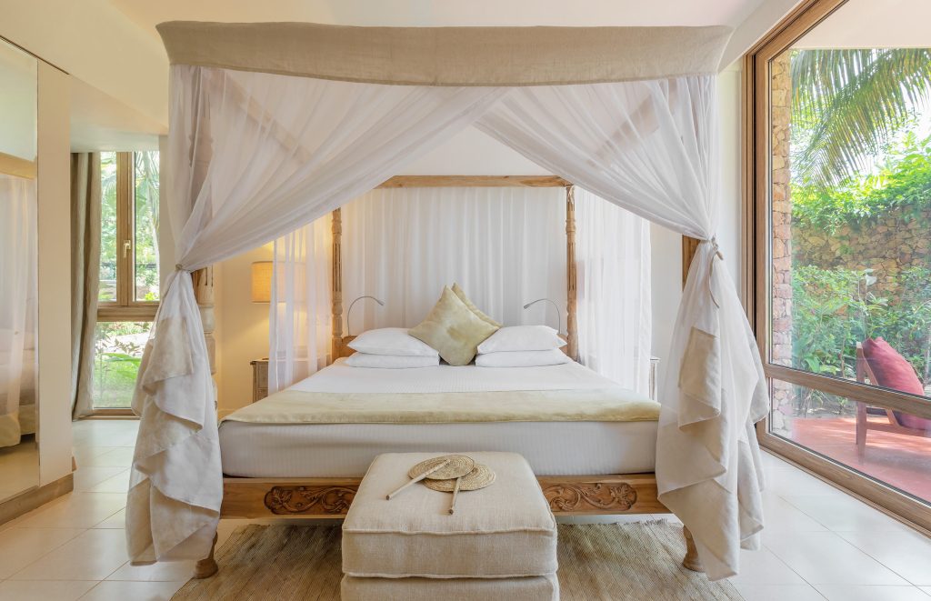 Schlafzimmer mit großem Himmelbett aus hellem Holz, weißen Vorhängen, Hocker mit Strohhut und großen Fenstern mit Gartenblick