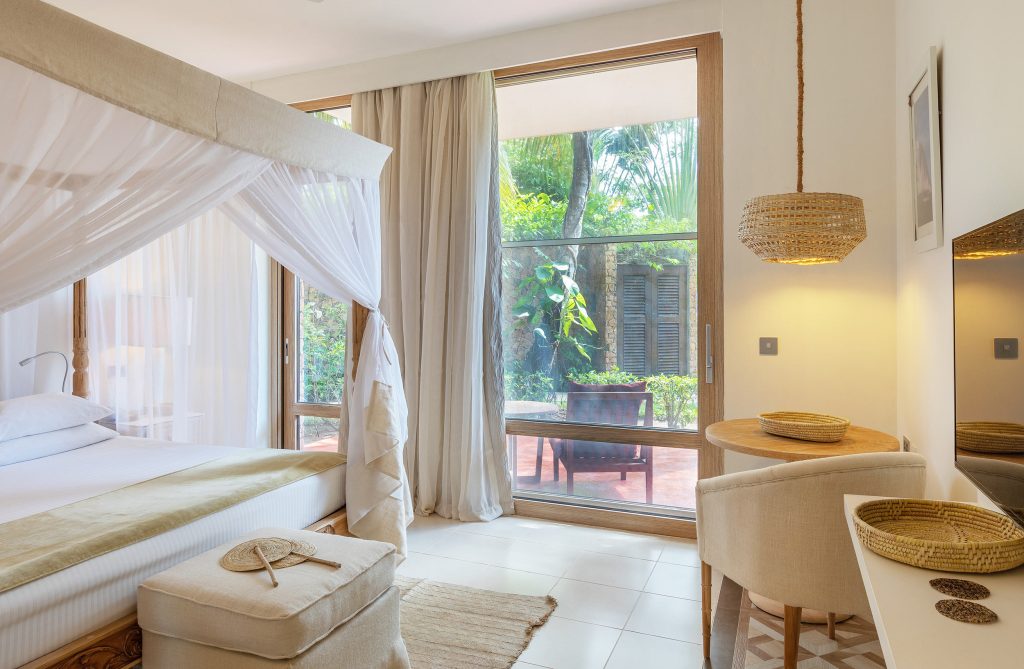 Hell beleuchtetes Hotelzimmer mit Himmelbett, Holzmöbeln und Blick auf Garten mit Steinmauern