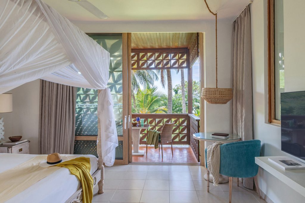 Hotelzimmer mit weißem Baldachinbett, Holzmöbeln und Balkon mit Blick auf tropische Vegetation in Sansibar