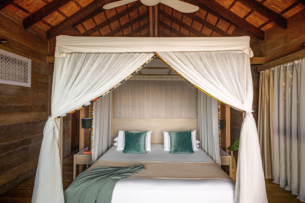 Schlafzimmer mit Himmelbett, weißen Vorhängen, dunklem Holz an Wänden und Decke sowie grünen Kissen im Melia Zanzibar