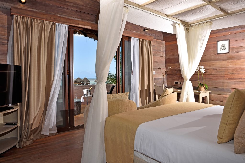 Hotelzimmer mit großem Bett, dunklen Holzwänden, Glasschiebetür zum Balkon mit Korbmöbeln und Meerblick in Sansibar