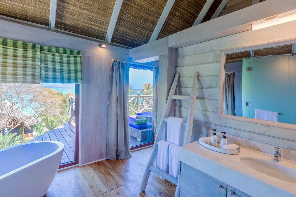 Badezimmer mit freistehender weißer Badewanne, großen Glasfenstern, Holzverkleidung und Blick auf tropische Pflanzen und Meer