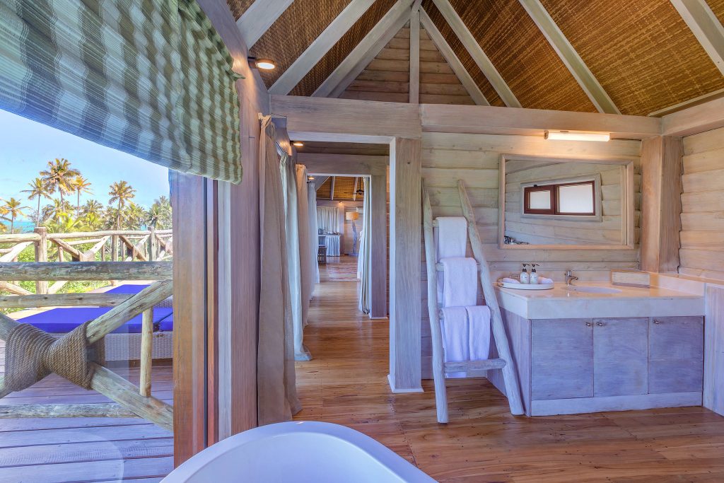 Hotelzimmer aus Holz mit großem Fenster, Waschtisch, Spiegel und Blick auf Terrasse mit Palmen in Sansibar