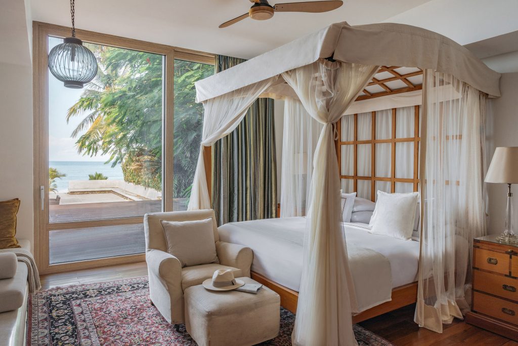 Hotelzimmer mit großem Himmelbett aus Holz, Sessel, Fußhocker, Tageslicht und Meerblick durch