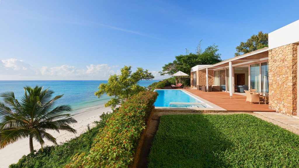 Moderner Pool mit Holzterrasse und flachem Steingebäude neben weißem Sandstrand bei Tageslicht