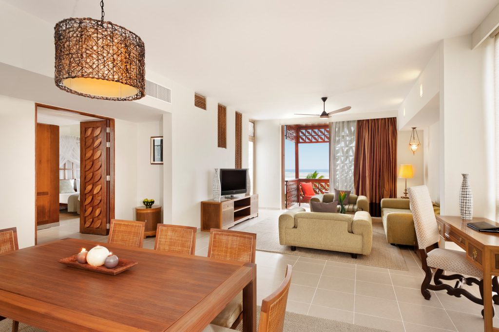 Offener Wohnbereich eines Hotelzimmers mit Holzesstisch, Korbstühlen, beigem Sofa und Blick auf Terrasse und Meer