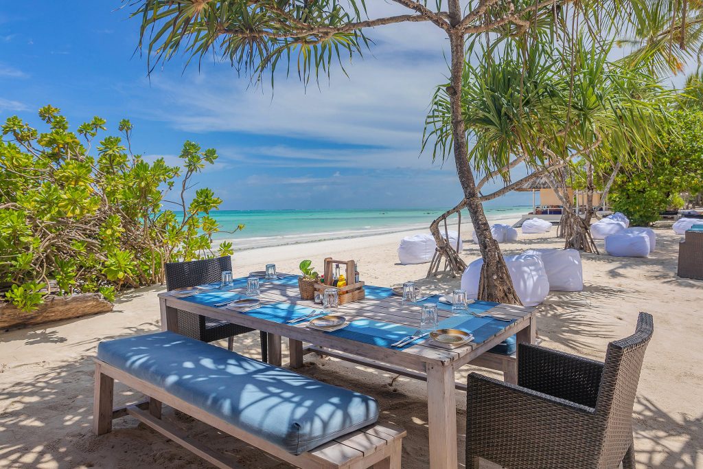 Strandbereich mit gedecktem Holztisch und Rattan-Stühlen, feinsandiger Strand und Palmen im Hintergrund