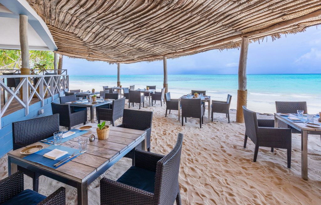 Offenes Strandrestaurant mit Holztischen und dunklen Rattanstühlen auf hellem Sand am Meer in Sansibar